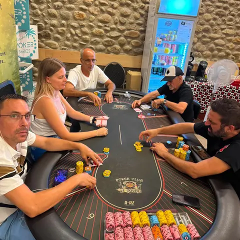 Une main en cours au Poker Club Le Soler : cinq joueurs autour du tapis aux couleurs du club, jetons empilés devant eux.