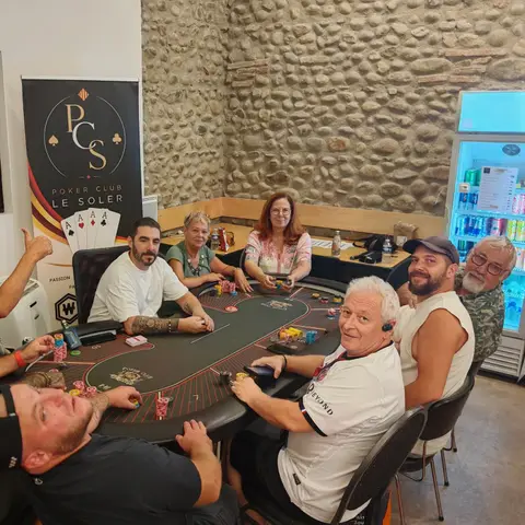 Une dizaine de joueurs souriants autour d'une table du Poker Club Le Soler, devant la bannière du club et le mur en galets de la salle.